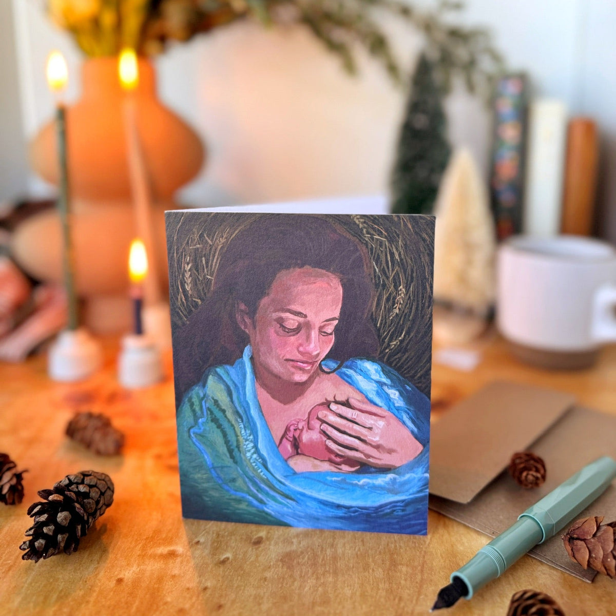 A greeting card featuring a painted portrait of a woman in a blue robe holding a baby, displayed on a wooden table with candles, pinecones, a pen, and other cozy decor in the background.