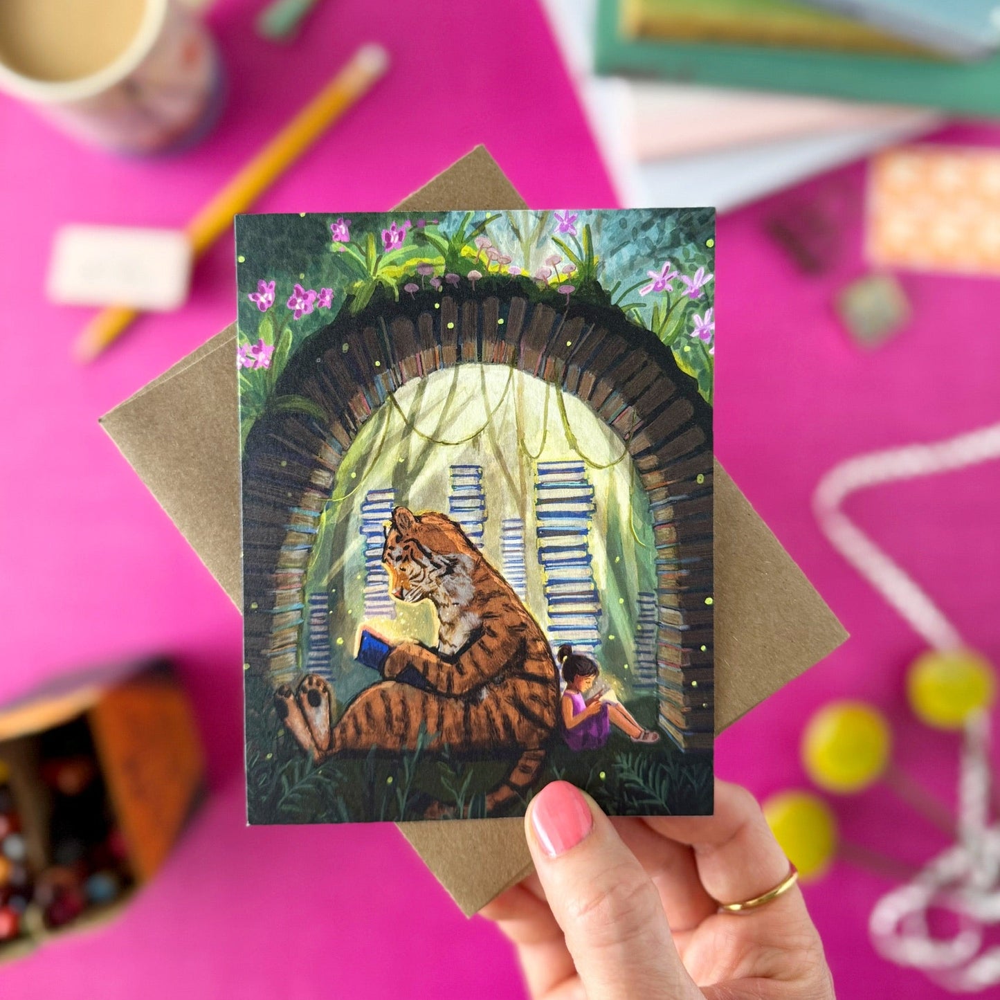 A hand holds a greeting card featuring an illustrated tiger and girl sitting inside a dark arched opening, surrounded by colorful books and stationery on a bright pink surface.