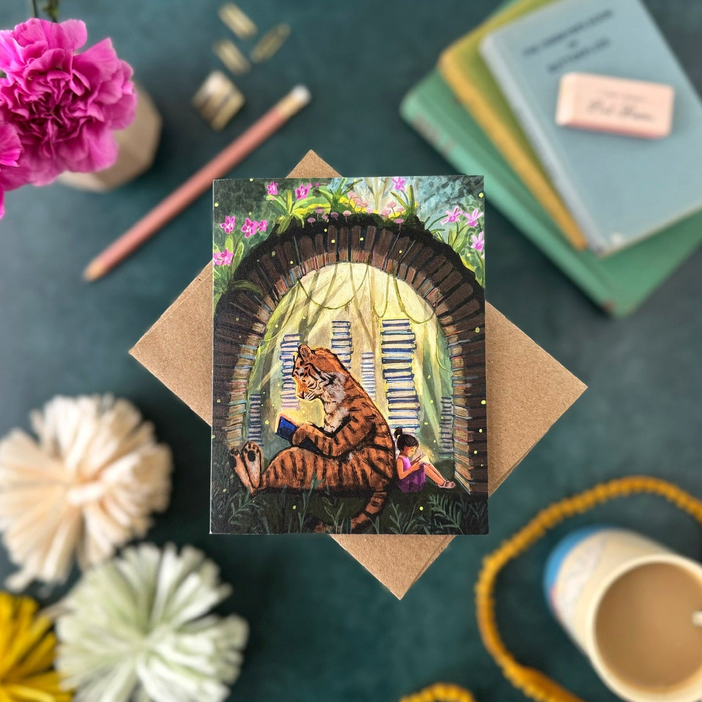 A square art print or card featuring an illustrated tiger and giftl reading sitting in front of stacks of books and under a book arch, styled in a flat lay on a teal surface with flowers, paper pom-poms, notebooks, a cup, and a string of beads.