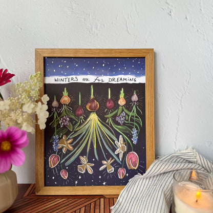 A framed art print featuring whimsical botanical illustrations of flowers and bulbs under a dark blue snowy sky with the text 'WINTERS are FOR DREAMING'. The print is framed in light wood and styled on a wooden surface next to a candle, a striped cloth, and flowers against a textured white wall.