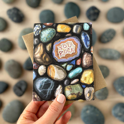 Hand holding a card with 'You Rock' design against a background of pebbles