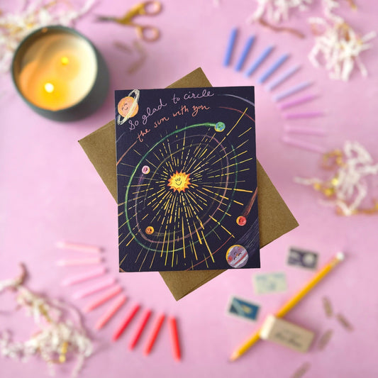 A greeting card featuring a friendly, colorful solar system illustration on a dark background, placed on top of a brown envelope. The card has handwritten text reading 'So glad to circle the sun with you.'