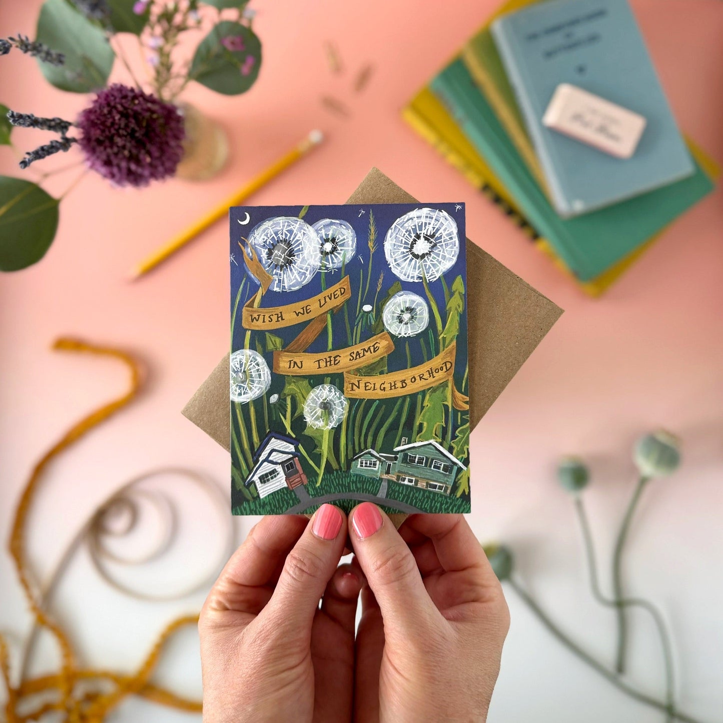 A hand holds a illustrated greeting card with a kraft envelope behind it on a pink tabletop. The card shows huge dandelions, small houses, and a banner reading 'Wish We lived in the Same Neighborhood."