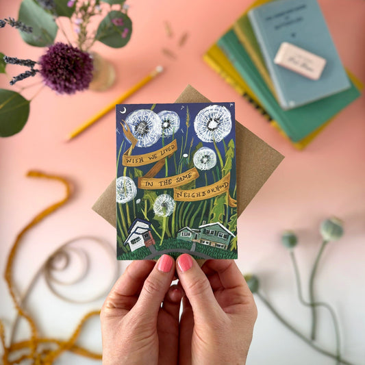 A hand holds a illustrated greeting card with a kraft envelope behind it on a pink tabletop. The card shows huge dandelions, small houses, and a banner reading 'Wish We lived in the Same Neighborhood."