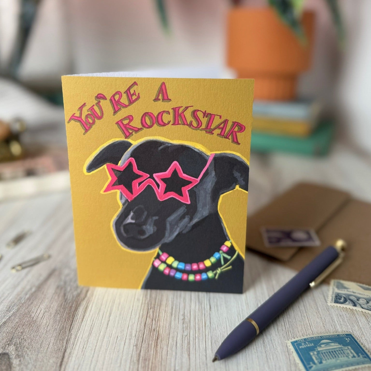 A greeting card featuring a black dog wearing pink star-shaped sunglasses and a colorful beaded necklace, with the text 'YOU'RE A ROCKSTAR' in pink. The card is displayed on a wooden desk next to a navy blue pen and postage stamps, with a blurred background showing a plant and books.